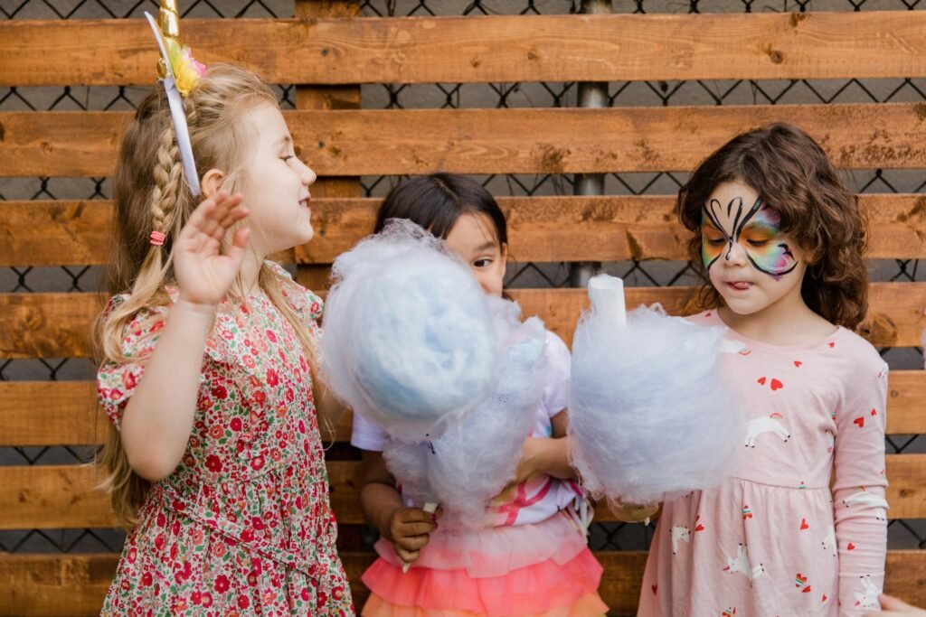 Three young girls enjoy cotton candy at an outdoor birthday party with face paint and unicorn headband.